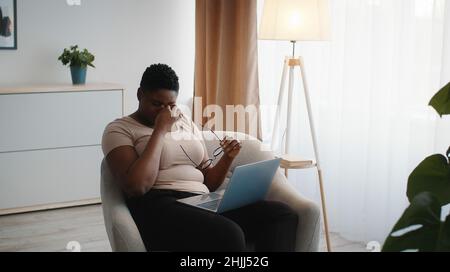 Erschöpfte afroamerikanische Frau, die die Brille auszieht und Nasenrücken reibt, Pause bei der Online-Arbeit auf dem Laptop macht Stockfoto