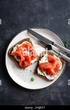 Köstlicher, gesalzener Lachs und Frischkäse-Roggen-Toast auf weißem Teller, schwarzer Schieferhintergrund, Draufsicht. Gesunde Vorspeise oder Snacks, offene Sandwiches Stockfoto