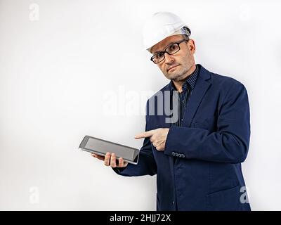 Ein Ingenieur in einem weißen, harten Helm, der einen Tablet-Computer hält, zeigt mit einem Finger darauf. Mann mittleren Alters in einer blauen Jacke. Isoliertes Porträt auf einem Stockfoto
