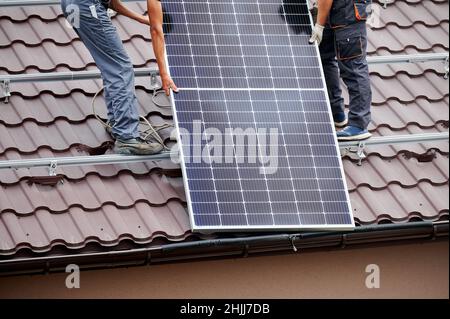 Männer Monteure heben Photovoltaik-Solarmodule auf dem Dach des Hauses. Beschnittene Ansicht von Elektrikern, die Solarpanel-System im Freien installieren. Konzept der alternativen und erneuerbaren Energien. Stockfoto