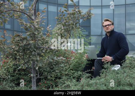 Mann, der im Park an einem Laptop arbeitet, außerhalb des Büros geschäftlich tätig ist, aus der Ferne lernt und arbeitet, freiberuflich tätig ist Stockfoto