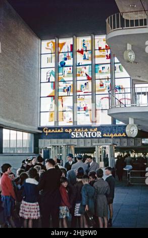 Passagiere, die am Bahnhof Eindhoven Centraal, Eindhoven, Nordbrabant, Niederlande um 1960 warten. Die Buntglaskunst (Glasapplikation) wurde vom niederländischen Künstler Lex Horn (1916–1968) angefertigt und 1955 an der Station installiert. Ein Neonschild wirbt für Senator-Zigarren. Der Hauptbahnhof Eindhoven ist der Hauptbahnhof der Stadt. Dieses Bild stammt von einer alten Amateur-Farbtransparenz bei schlechten Lichtverhältnissen – einem Vintage-Foto aus dem Jahr 1950s/1960s. Stockfoto