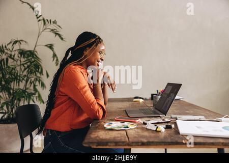 Glückliche Grafikdesignerin, die an einem virtuellen Meeting teilnahm, während sie an ihrem Schreibtisch saß. Kreative junge Frau, die mit einem Laptop Videoanruf bei ihren Kunden führt. Weiblich Fr. Stockfoto