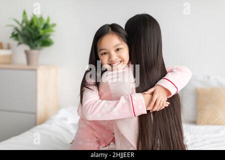 Zufriedene glückliche chinesische Teenager-Mädchen umarmte junge Dame auf dem Bett im Schlafzimmer Innenraum, freien Platz Stockfoto