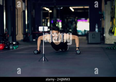 Athletischer Arabischer Mann, Der Dumbbell-Liegestütze Macht, Während Er Inhalte Auf Einer Smartphone-Kamera Aufzeichte Stockfoto