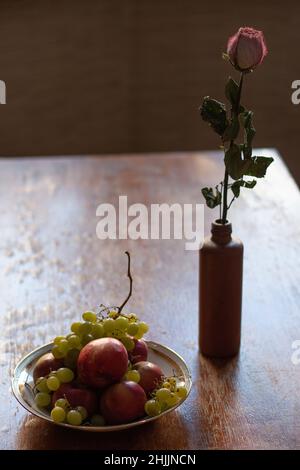 Trockene Rosenblüte in einem Tongefäß, neben einer Schale mit Früchten, Trauben und Äpfeln, auf einem roten Holztisch, Stillleben, vertikales Foto Stockfoto