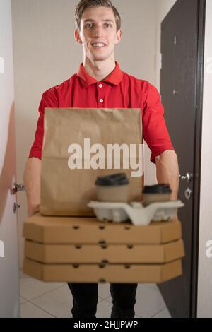 Pizza- und Kaffeelieferung, ein kaukasischer Mann in roter Uniform liefert Pizza, wickelt Pizzakisten ab, gibt dem Kunden vor der Tür Essen. Stockfoto