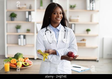 Porträt einer fröhlichen jungen schwarzen Ernährungswissenschaftlerin mit Apfel und Klebeband, die Obst und Gemüse empfiehlt, Freiraum Stockfoto