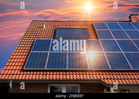 Fotocollage von Solarzellen, Photovoltaik auf dem roten Dach eines Hauses und einem schönen Himmel mit untergehenden Sonne. Alternative Stromversorgung. Stockfoto