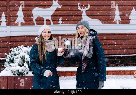 Zwei junge, fröhlich lächelnde Frauen im Schneefall werfen Schneebälle auf die Kamera. Stockfoto