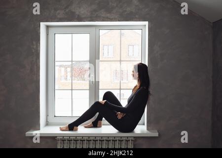 Eine Frau in Sportkleidung sitzt auf der Fensterbank und blickt aus dem Fenster auf den Schneefall Stockfoto