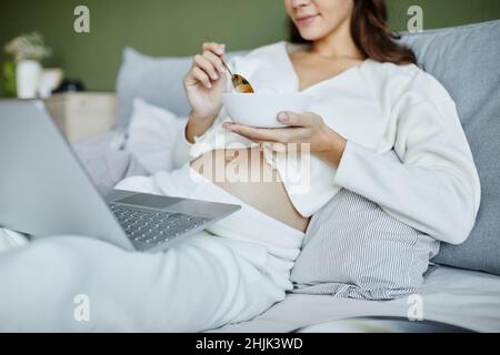 Junge Schwangere, die mit einem Laptop auf den Knien auf dem Bett sitzt und beim Frühstück im Schlafzimmer etwas beobachtet Stockfoto