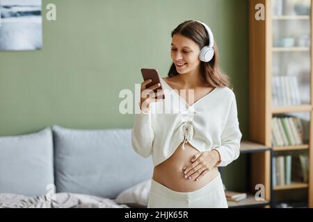 Junge Schwangerin mit kabellosen Kopfhörern, die die Musik auf ihrem Mobiltelefon genießen und im Zimmer tanzen Stockfoto
