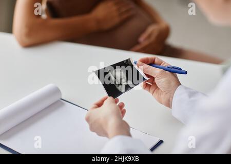 Nahaufnahme eines Arztes, der das Ultraschallbild des Embryos untersucht, das am Tisch sitzt und während ihres Besuchs mit einer Schwangeren spricht Stockfoto
