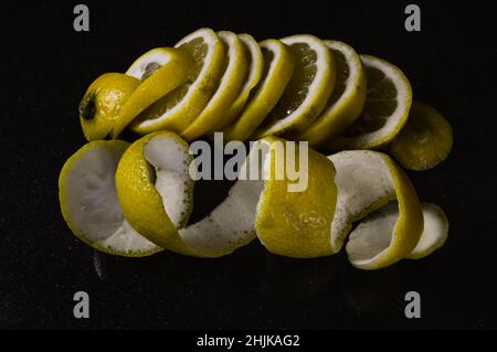 Zitronenscheiben und ganze Zitronenschale in einer Spirale auf einem glitzernden schwarzen Glashintergrund zusammengerollt Stockfoto