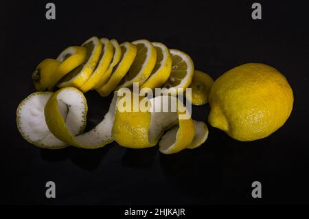 Zitronenscheiben und ganze Zitronenschale in einer Spirale gekräuselt und eine ganze Zitrone auf einem glitzernden schwarzen Glashintergrund Stockfoto