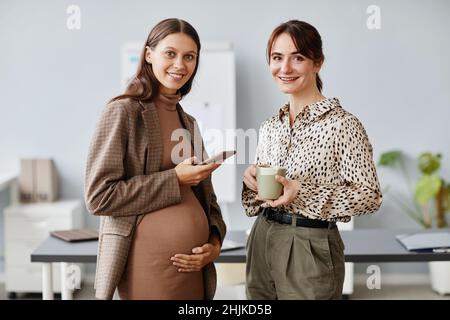 Porträt einer Schwangeren Geschäftsfrau mit Mobiltelefon lächelt vor der Kamera zusammen mit ihrer Kollegin bei einer Tasse Kaffee, sie stehen im Büro Stockfoto
