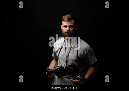 Professioneller hübscher bärtiger männlicher Fotograf mit einer Reihe von Vintage-Fotokameras im Fotostudio, isoliert auf schwarzem Hintergrund. Stockfoto