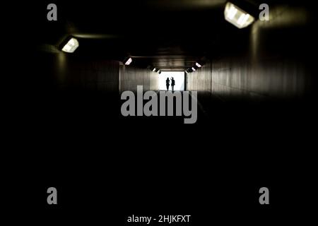 Zwei Silhouetten am Ende des Tunnels. Licht in der Ferne. Figuren von kleinen Frauen in kontrastierendem Licht. Lampen in langer Unterführung. Stockfoto