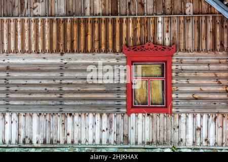 Ein Vintage-Fenster mit karmesinrotem Zierrahmen in brauner Holzwand. Altes Wohnhaus des russischen Bauern in Lettland. Stockfoto