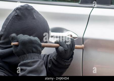 Räuber Mann in schwarzer Kapuzenjacke mit einem Brecheisen, um Schloss zu brechen und ein Fahrzeug zu stehlen. Autodieb oder Diebstahl für Versicherungskonzept. Stockfoto