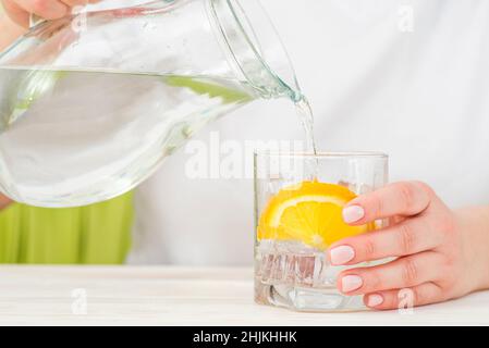 Weibliche Hände gießen Wasser aus dem Dekanter in einen Glasbecher mit Zitrone und Eis. Gesundheit und Ernährung Konzept. Durst an einem heißen Tag löschen. Stockfoto