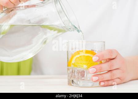 Weibliche Hände gießen Wasser aus dem Dekanter in einen Glasbecher mit Zitrone und Eis. Gesundheit und Ernährung Konzept. Durst an einem heißen Tag löschen. Stockfoto