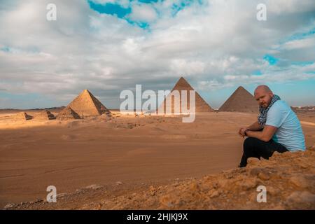 Mann, der auf den sandigen Wüstendünen sitzt und vor den großen Pyramiden von gizeh posiert. Reisen ägypten im Winter, Touristen posieren für ein Bild Stockfoto