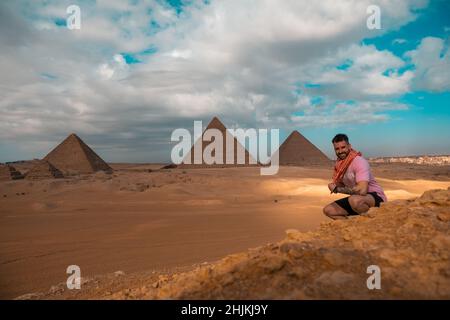 Mann, der auf den sandigen Wüstendünen sitzt und vor den großen Pyramiden von gizeh posiert. Reisen ägypten im Winter, Touristen posieren für ein Bild Stockfoto