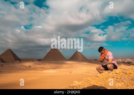 Mann, der auf den sandigen Wüstendünen sitzt und vor den großen Pyramiden von gizeh posiert. Reisen ägypten im Winter, Touristen posieren für ein Bild Stockfoto