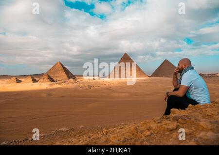 Mann, der auf den sandigen Wüstendünen sitzt und vor den großen Pyramiden von gizeh posiert. Reisen ägypten im Winter, Touristen posieren für ein Bild Stockfoto