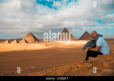 Mann, der auf den sandigen Wüstendünen sitzt und vor den großen Pyramiden von gizeh posiert. Reisen ägypten im Winter, Touristen posieren für ein Bild Stockfoto