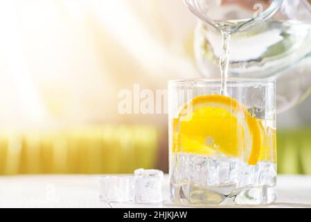 Weibliche Hände gießen Wasser aus dem Dekanter in einen Glasbecher mit Zitrone und Eis. Gesundheit und Ernährung Konzept. Durst an einem heißen Tag löschen. Stockfoto