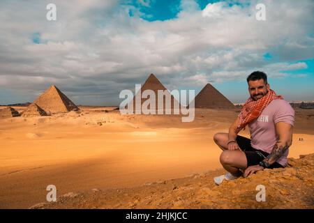 Mann, der auf den sandigen Wüstendünen sitzt und vor den großen Pyramiden von gizeh posiert. Reisen ägypten im Winter, Touristen posieren für ein Bild Stockfoto