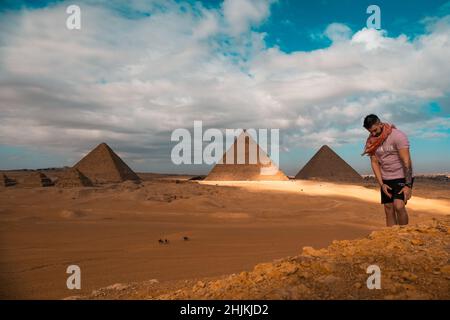 Mann, der auf den sandigen Wüstendünen sitzt und vor den großen Pyramiden von gizeh posiert. Reisen ägypten im Winter, Touristen posieren für ein Bild Stockfoto