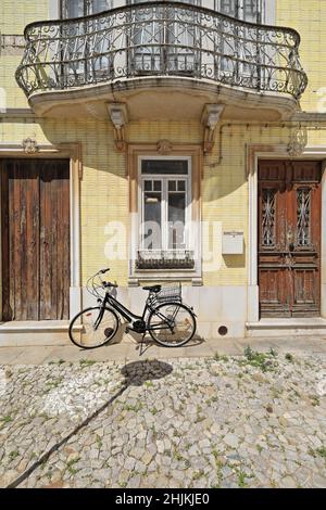 Fassade mit gelben Kacheln, neoklassizistisches Stadthaus, geplatzte Holztüren, Metallgeländer, Balkon, schwarzes Fahrrad. Tavira-Portugal-068 Stockfoto