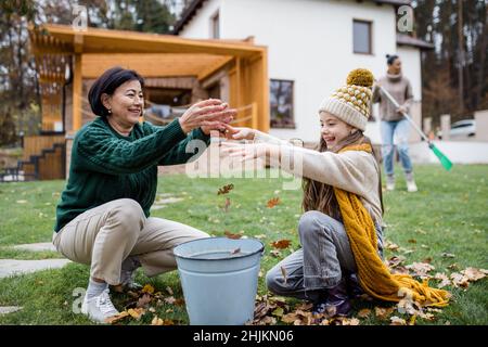 Glückliches kleines Mädchen mit Großmutter, die Blätter aufhob und sie im Herbst in den Eimer im Garten legte Stockfoto