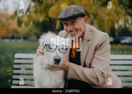 Glücklicher älterer Mann, der die Kamera ansah und seinen Hund mit einer Brille auf der Bank im Freien in der Stadt umarmte. Stockfoto