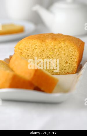 Selbstgebackener Kuchen für den Nachmittagstee, in Scheiben geschnitten auf einem weißen rechteckigen Teller mit weißer Teekane und Tasse im Hintergrund Stockfoto
