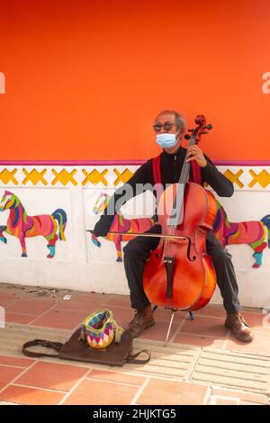 Guatape, Antioquia, Kolumbien - 8 2021. Dezember: Der alte, in Schwarz gekleidete braune Mann mit roten Strapsen und Sonnenbrillen spielt auf einer Straße das Cello Stockfoto