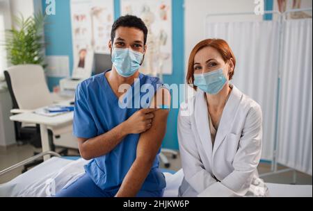 Arzt mit einem Kollegen, der einen medizinischen Gesichtsmast trägt, die Kamera anschaut und nach dem Impfstoff einen Verband zeigt Stockfoto