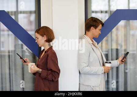 Seitenansicht eines Porträts von zwei Frauen, die Smartphones während der Büropause benutzen und sich zurück an den Rücken stellen, um Platz zu kopieren Stockfoto