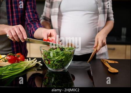 Konzentrieren Sie sich auf männliche Hände, die ein Küchenmesser halten und geschnittene Tomaten in eine Schüssel mit Salatblättern und Grüns auf dem Hintergrund seines schwangeren wi krönen Stockfoto