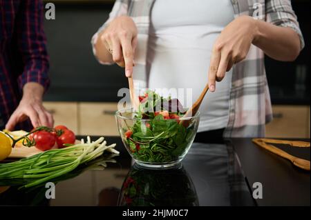 Konzentrieren Sie sich auf die Hände einer Schwangeren, die Holzlöffel halten und Zutaten in einer Glasschüssel mischen, um im Kitc köstlichen gesunden Salat zum Abendessen zuzubereiten Stockfoto