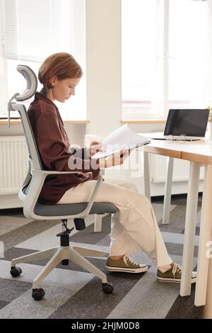 Seitenansicht Ganzkörperportrait mit tätowierten jungen Frauen, die Dokumente lesen, während sie am Schreibtisch in einem modernen, hellen Büro sitzen Stockfoto