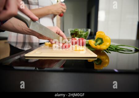 Nahaufnahme der Hände eines Mannes, die Tomaten und gelbe Paprika auf einem Schneidebrett auf dem Hintergrund einer Schwangeren schneiden, die einen Zweig mit frischem Toma hält Stockfoto