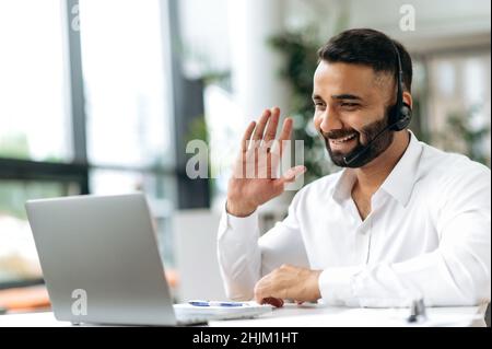 Gesellig attraktiver, intelligenter indischer Unternehmer, Geschäftsmann, Verkaufsmanager, Händler, mit Kopfhörern sitzen am Tisch im Büro, kommuniziert per Videoanruf mit Kollegen oder Kunden, grüßt, lächelt Stockfoto