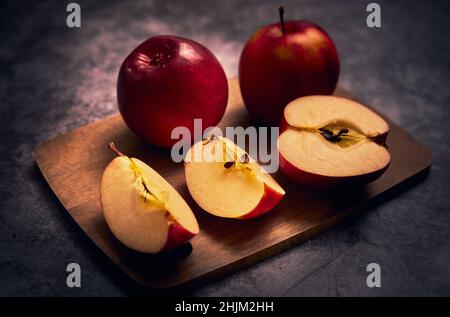 Apfelbrett. Äpfel stehen auf einem Schneidebrett und ein Apfel wird in mehrere Stücke geschnitten und die ganze Komposition auf einem dunklen Hintergrund Stockfoto