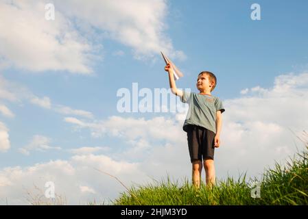 Kleiner Junge startet ein Papierflugzeug in die Luft. Das Kind startet eine Papierebene. Glückliches Kind, das mit Papierflugzeug spielt. Stockfoto