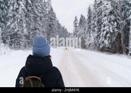 Frau Reisende Blick in die Ferne auf sich bewegende Auto, während auf verschneite Straße im Winterwald stehen Stockfoto